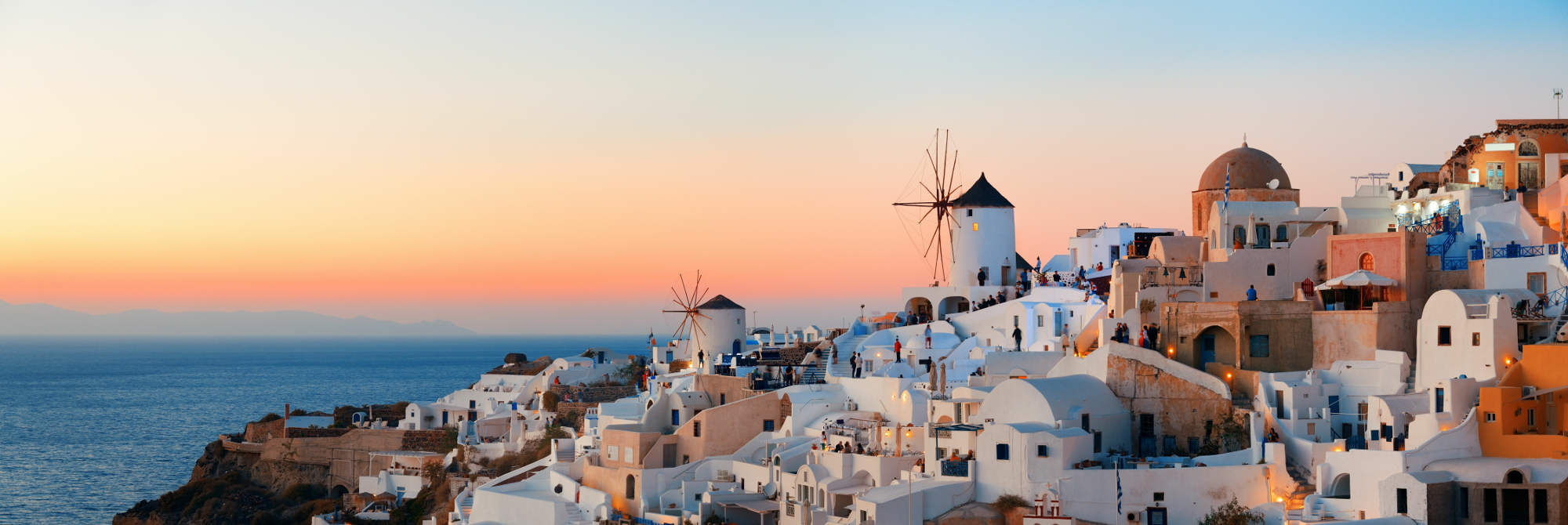 Weiße Häuser mit blauen Dächern und zwei Windmühlen in Oia auf Santorini bei Sonnenuntergang mit Blick auf das Meer.
